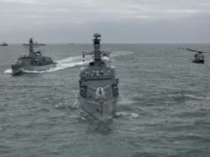 British frigate and helicopter fire torpedo in the Atlantic Three gray military ships sail in formation on the open sea, with a helicopter hovering to the right.