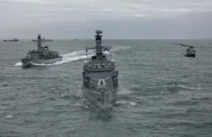 Three gray military ships sail in formation on the open sea, with a helicopter hovering to the right.