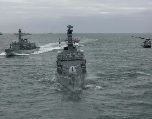 British frigate and helicopter fire torpedo in the Atlantic Three gray military ships sail in formation on the open sea, with a helicopter hovering to the right.