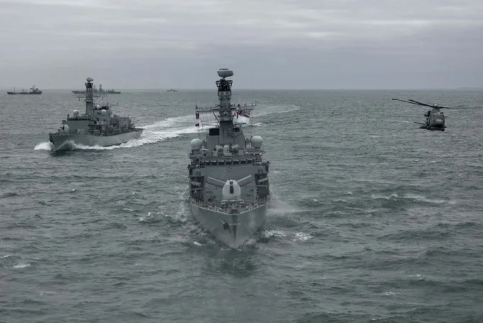 Three gray military ships sail in formation on the open sea, with a helicopter hovering to the right.