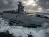 Gray military destroyer with hull marked X11 cuts through rough, stormy seas as spray and dark clouds loom overhead.