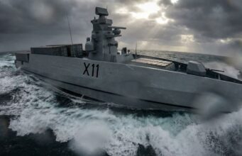 Gray military destroyer with hull marked X11 cuts through rough, stormy seas as spray and dark clouds loom overhead.
