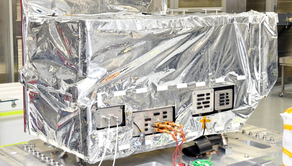 Reflective-wrapped satellite instrument in a cleanroom, with exposed control panels and orange cables visible.