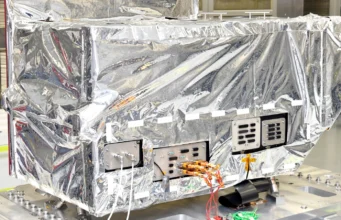 Reflective-wrapped satellite instrument in a cleanroom, with exposed control panels and orange cables visible.
