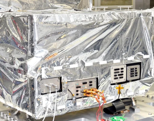 Reflective-wrapped satellite instrument in a cleanroom, with exposed control panels and orange cables visible.