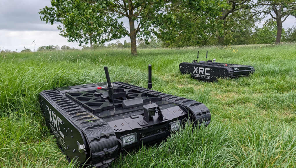 PXL_20260520_150643676 Two black tracked robots with antennas sit in a tall-grass field, one foreground and one in the background, labeled XRC Robotics.