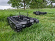 Two black tracked robots with antennas sit in a tall-grass field, one foreground and one in the background, labeled XRC Robotics.