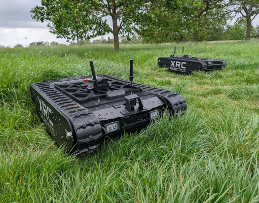 Two black tracked robots with antennas sit in a tall-grass field, one foreground and one in the background, labeled XRC Robotics.