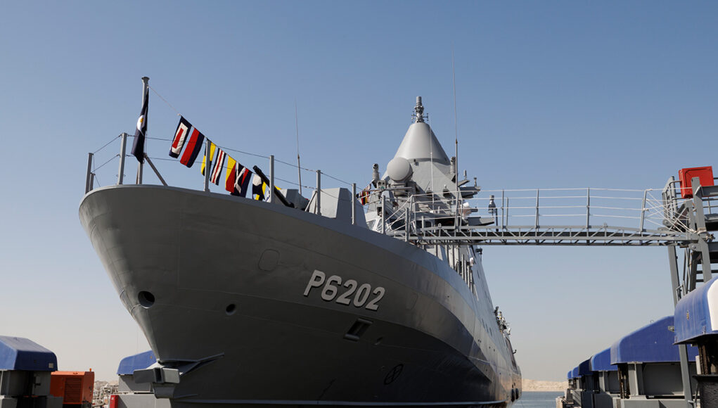 Large gray naval vessel named P6202 docked at a pier, with signal flags along the bow and a gangway extending to shore.