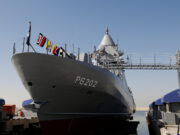 Large gray naval vessel named P6202 docked at a pier, with signal flags along the bow and a gangway extending to shore.