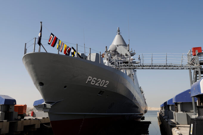 Large gray naval vessel named P6202 docked at a pier, with signal flags along the bow and a gangway extending to shore.