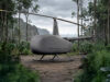 Grey autonomous helicopter drone with a two-bladed rotor on a forest path, rugged landing skids, surrounded by dense jungle.