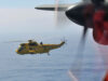 Yellow RAF search-and-rescue helicopter flying low over the ocean, seen through the window of another aircraft with a blurred foreground propeller.