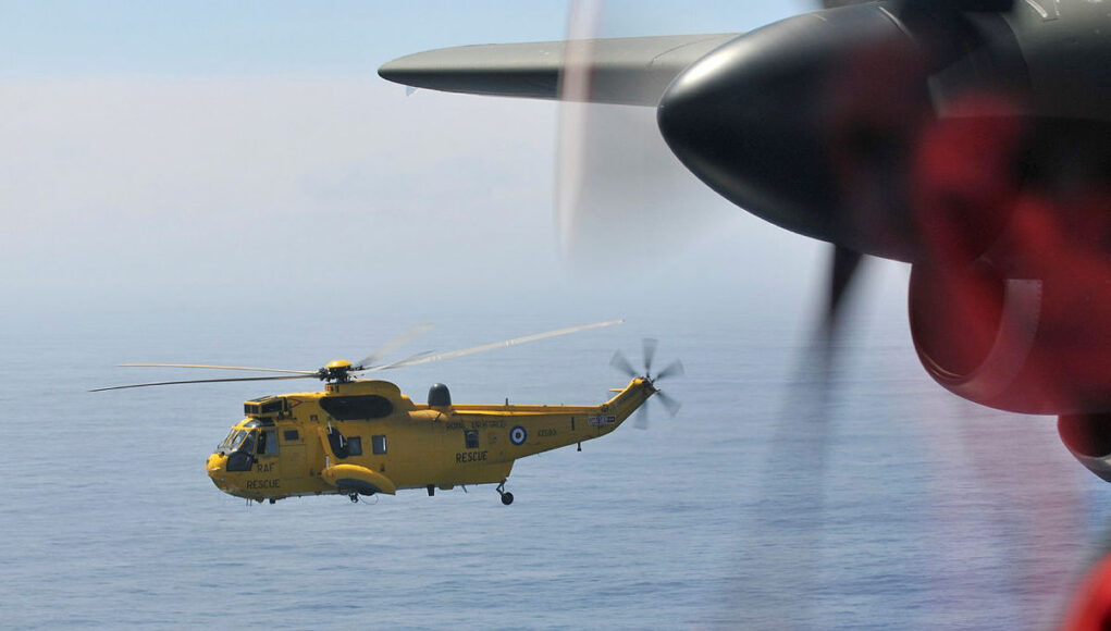 Yellow RAF search-and-rescue helicopter flying low over the ocean, seen through the window of another aircraft with a blurred foreground propeller.