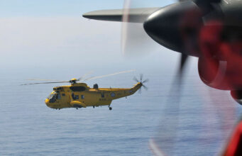 Yellow RAF search-and-rescue helicopter flying low over the ocean, seen through the window of another aircraft with a blurred foreground propeller.