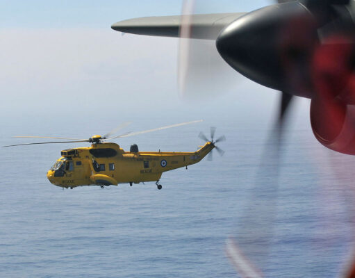 Yellow RAF search-and-rescue helicopter flying low over the ocean, seen through the window of another aircraft with a blurred foreground propeller.