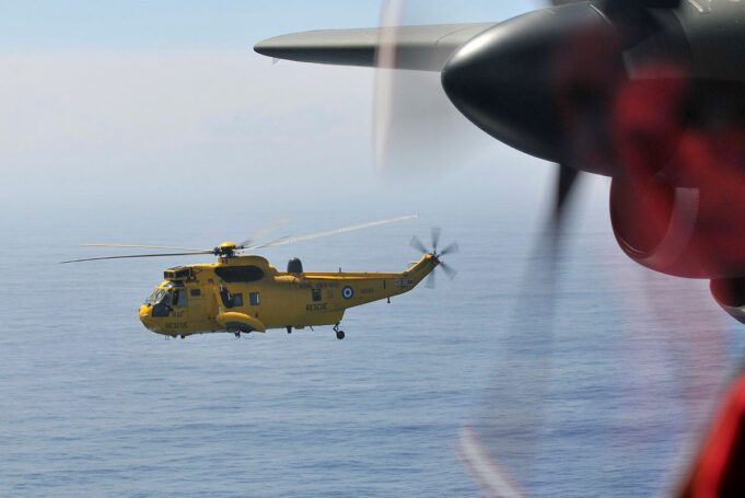 Yellow RAF search-and-rescue helicopter flying low over the ocean, seen through the window of another aircraft with a blurred foreground propeller.