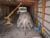 Large metal V-shaped framework (ship hull) inside a hangar-like workshop, lifted by a yellow crane overhead.