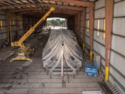 Large metal V-shaped framework (ship hull) inside a hangar-like workshop, lifted by a yellow crane overhead.