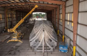 Large metal V-shaped framework (ship hull) inside a hangar-like workshop, lifted by a yellow crane overhead.