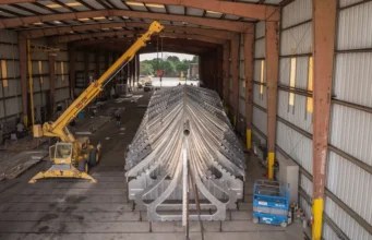 Large metal V-shaped framework (ship hull) inside a hangar-like workshop, lifted by a yellow crane overhead.