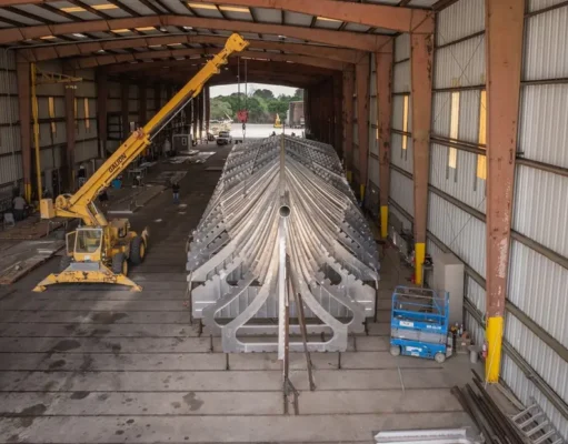 Large metal V-shaped framework (ship hull) inside a hangar-like workshop, lifted by a yellow crane overhead.
