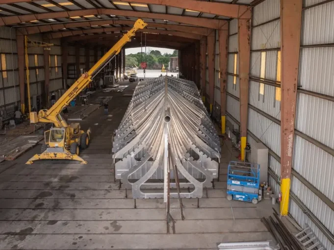 Large metal V-shaped framework (ship hull) inside a hangar-like workshop, lifted by a yellow crane overhead.