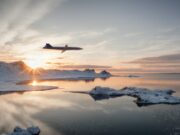 Jet gliding over a snow-covered coastline at sunset, reflected in the calm icy water below.