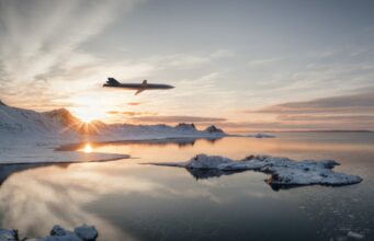 Jet gliding over a snow-covered coastline at sunset, reflected in the calm icy water below.