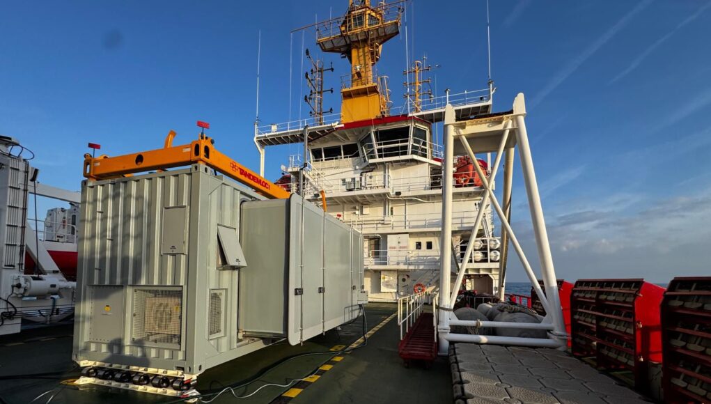 Royal-Navy-achieves-milestone-with-first-sea-trials-of-NavyPODS Deck of a large support vessel with a gray containerized generator and an orange crane, against a blue sky and ocean.”