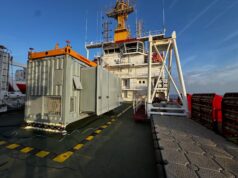 Deck of a large support vessel with a gray containerized generator and an orange crane, against a blue sky and ocean.&rdquo;