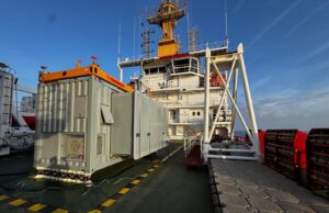 Deck of a large support vessel with a gray containerized generator and an orange crane, against a blue sky and ocean.&rdquo;
