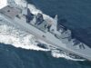Modern gray destroyer cruising through open sea, bow to the right, leaving a frothy wake behind.