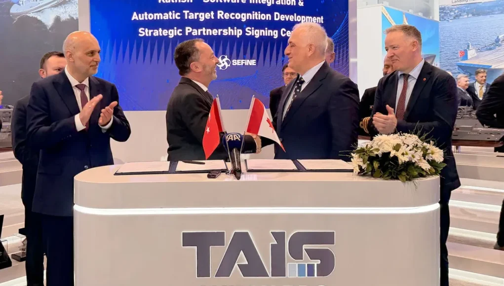 Two men in suits shake hands across a signing desk with Turkish and Georgian flags; colleagues applaud behind a TAIS booth.