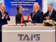 Two men in suits shake hands across a signing desk with Turkish and Georgian flags; colleagues applaud behind a TAIS booth.