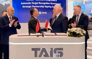 Two men in suits shake hands across a signing desk with Turkish and Georgian flags; colleagues applaud behind a TAIS booth.
