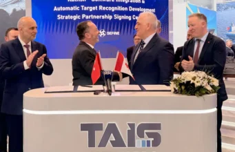 Two men in suits shake hands across a signing desk with Turkish and Georgian flags; colleagues applaud behind a TAIS booth.