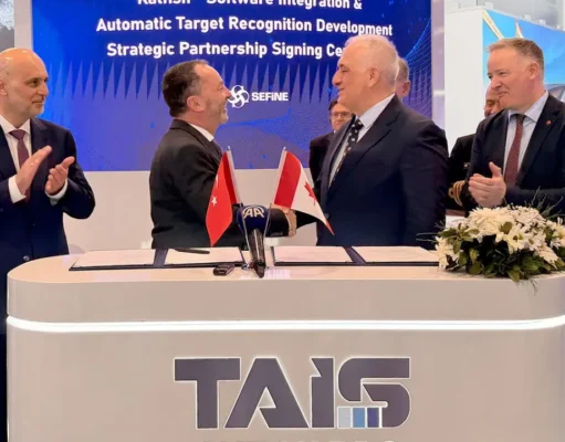 Two men in suits shake hands across a signing desk with Turkish and Georgian flags; colleagues applaud behind a TAIS booth.