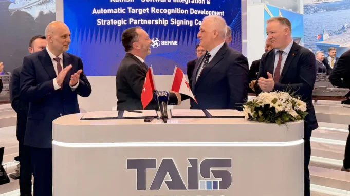 Two men in suits shake hands across a signing desk with Turkish and Georgian flags; colleagues applaud behind a TAIS booth.