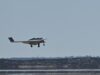 Small white single-engine airplane with landing gear down flying low over a flat coastal landscape and water.