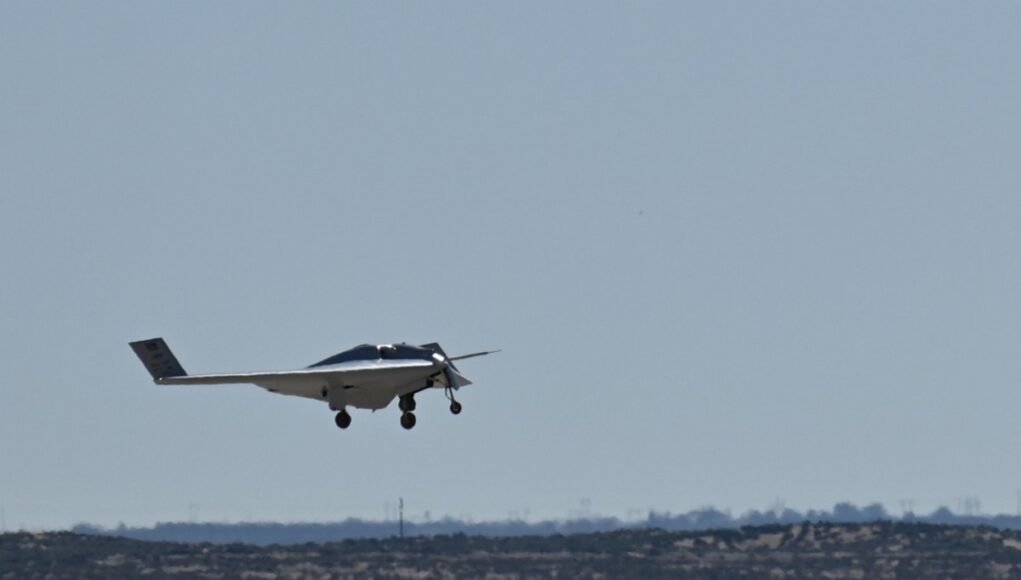 Small white single-engine airplane with landing gear down flying low over a flat coastal landscape and water.