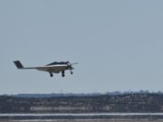 Small white single-engine airplane with landing gear down flying low over a flat coastal landscape and water.