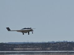 Small white single-engine airplane with landing gear down flying low over a flat coastal landscape and water.