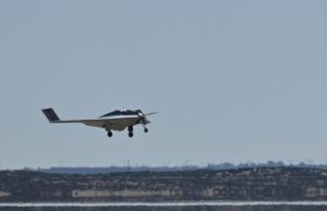 Northrop Grumman hybrid electric drone begins flight tests Small white single-engine airplane with landing gear down flying low over a flat coastal landscape and water.