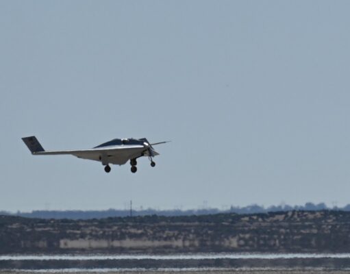 Small white single-engine airplane with landing gear down flying low over a flat coastal landscape and water.