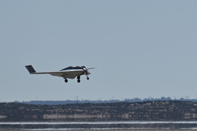 Small white single-engine airplane with landing gear down flying low over a flat coastal landscape and water.