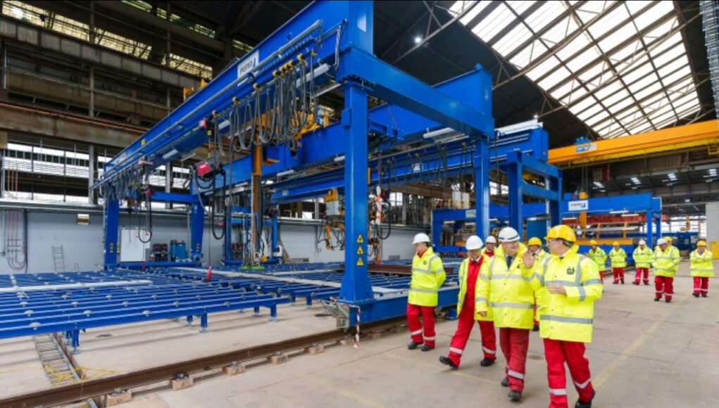 Group of workers in yellow high-visibility jackets and white hard hats walking beside a large blue overhead crane in an industrial factory setting with rails on the floor.