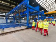 Group of workers in yellow high-visibility jackets and white hard hats walking beside a large blue overhead crane in an industrial factory setting with rails on the floor.