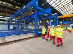 Group of workers in yellow high-visibility jackets and white hard hats walking beside a large blue overhead crane in an industrial factory setting with rails on the floor.