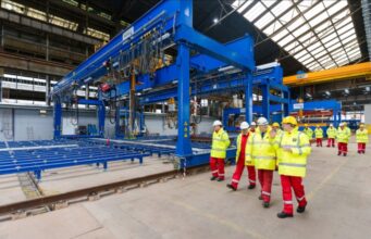 Group of workers in yellow high-visibility jackets and white hard hats walking beside a large blue overhead crane in an industrial factory setting with rails on the floor.